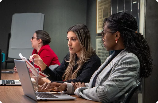 Three colleagues working on laptops side by side at a boardroom table during a meeting