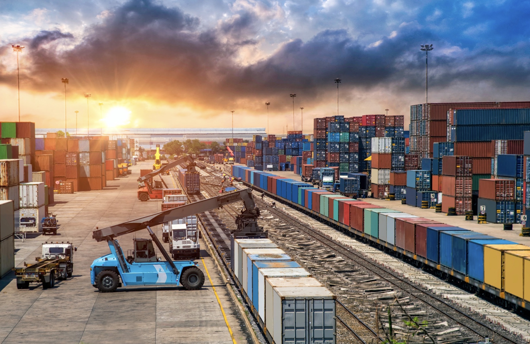 Terminal de conteneurs au coucher du soleil, avec un chariot gerbeur à côté d’un train de marchandises chargé de conteneurs maritimes
